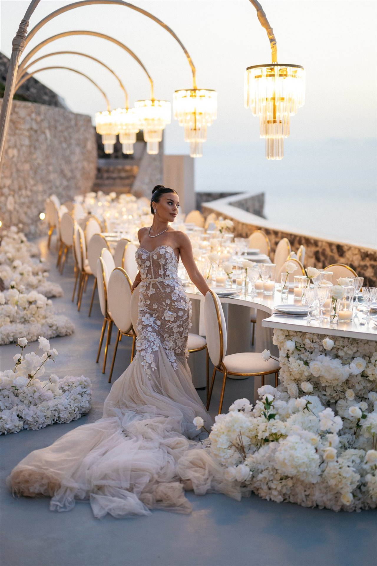 Bride beside a long floral dinner table above the Santorini caldera at sunset